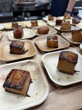 A row of small plates featuring beautifully caramelized pork belly squares garnished with herbs, served as appetizers at a Fork and Coin event. The image complements the caption by showcasing the carefully plated food from the exclusive gathering.