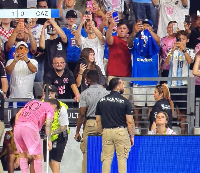 A stadium scene with a scoreboard showing '1 0 CAZ', where fans and security guards are visible, with a player in a pink uniform appearing injured.