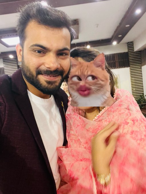 A selfie of a smiling bearded man in a maroon blazer standing next to a woman in a pink outfit whose face is edited with a kissing cat filter.