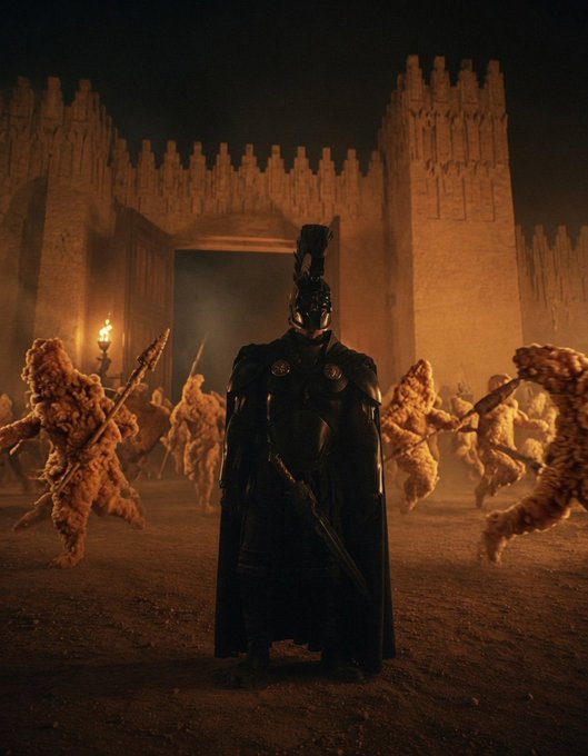 A dark cinematic scene shows a figure in black armor and cape holding a sword, standing before a glowing castle gate as dozens of golden, popcorn-like creatures with weapons swarm around them.