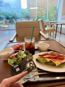 A hand displays a dark Visa credit card on a wooden tray, paired with an iced coffee, salad, and a BLT sandwich in a bright, plant-filled café. The post's caption roughly translates to: "This kind of setup is ideal, but the physical card's presence isn't required—please don't look too closely since the card is worn from overuse lol," humorously explaining why the card is held up to avoid showing its scuffed surface on the table.