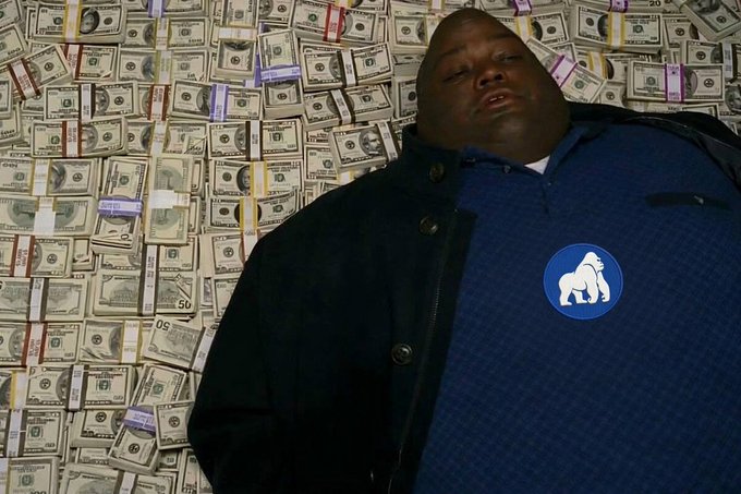 A man in a blue shirt with a white buffalo logo stands in front of a wall covered in stacks of US dollar bills.