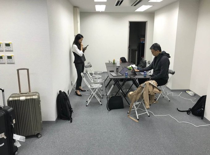 Two people work on laptops at a folding table in a small, plain office room, with luggage visible in the foreground.