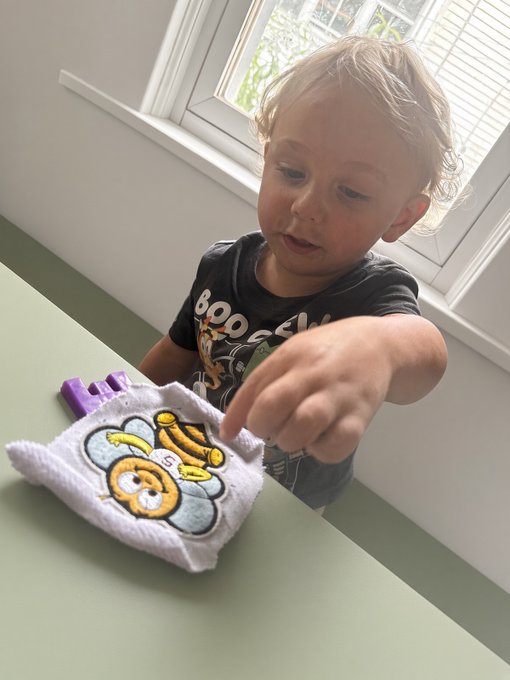 A young blonde child in a dark "BOO" t-shirt sits at a light green table, reaching out toward a white cloth hair scrunchie with a bear design and a purple claw clip.