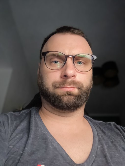 A selfie of a bearded man wearing glasses and a gray V-neck shirt.