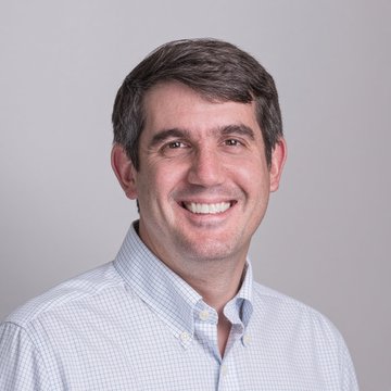A professional headshot portrait of a smiling middle-aged man with short brown hair, wearing a light blue checkered button-up shirt against a grey background.