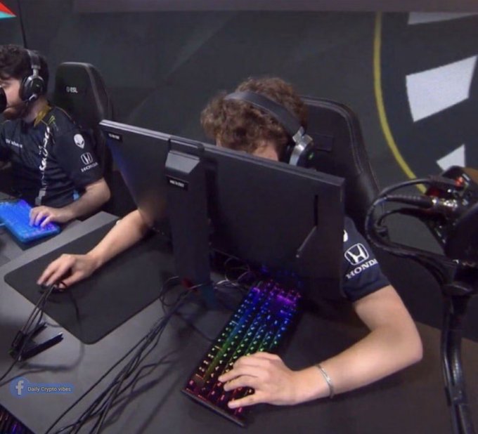 The image shows two people sitting at a table with computer monitors, one of them wearing headphones. They are using keyboards and mice while playing video games or working on computers. The background features a black wall with a logo that reads "Honda".