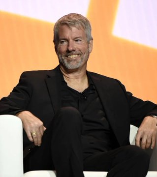 A photograph of a smiling bearded man in a black suit jacket and dark shirt, seated in a white chair against an orange background, appearing to be at a conference or panel.