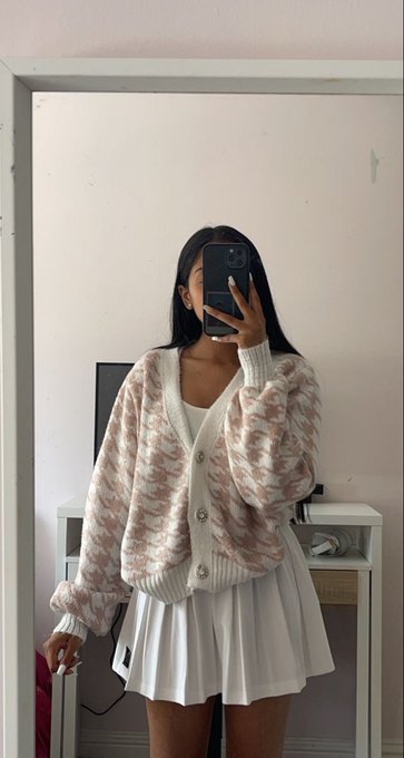 A mirror selfie of a young woman wearing a cream-colored patterned cardigan over a white pleated skirt, holding a phone that obscures her face in a simple room.