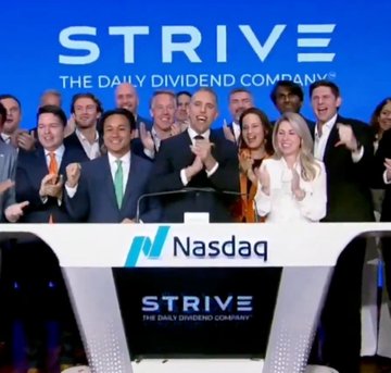 The image shows a group of people standing around a podium or stage area, possibly participating in an event like a presentation or award ceremony. On the podium, there's a sign that says "STRIVE" written in blue letters at the top. In front of this sign, there is a banner displaying the word "bitcoin". It appears to be a public announcement, perhaps related to digital currency or an innovation by a company named "STRIVE", which has gained significant attention recently. The people are smiling and clapping, indicating a positive atmosphere and excitement about something happening in the image.