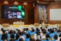 An audience of people in blue uniforms sits facing a stage with a presentation screen.