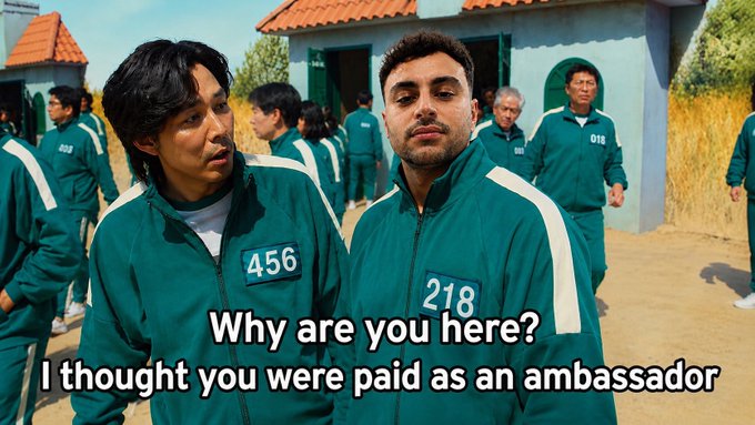 Two men in green tracksuits with number patches (456 and 218) stand in what appears to be a Squid Game scene, with text overlaid: 'Why are you here? I thought you were paid as an ambassador'.