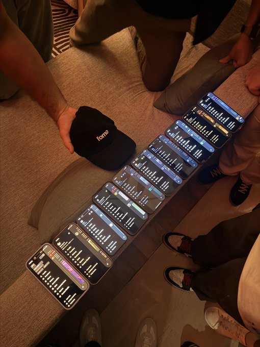 An overhead photo of a group of friends standing around a couch, each holding a smartphone displaying what appears to be a stock or crypto trading app, while one person holds up a black 'Fomo' cap.