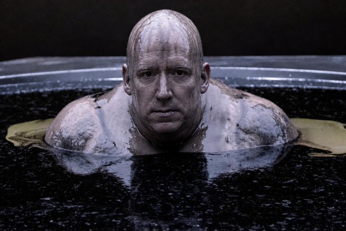 A bald man emerges from a dark liquid pool, his skin glistening with the fluid in a dramatic, cinematic shot.