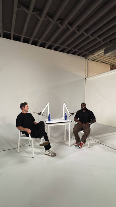 Two men sit across from each other at a small table with microphones in a white studio space, appearing to record a podcast.