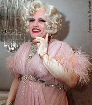 A blonde woman in elaborate theatrical makeup and a pink feathered dress poses playfully with her finger near her face, with a credit reading 'Spencer A. Burmett'.