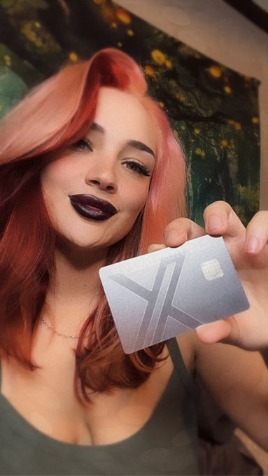 A portrait of a young woman with pink hair and dark lipstick holding up a frosted card with an "X" logo, likely the X (formerly Twitter) promotional card.