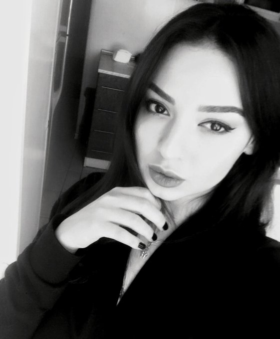 A black and white selfie of a young woman with long dark hair, dramatic eyeliner, and dark lipstick, resting her hand near her face.