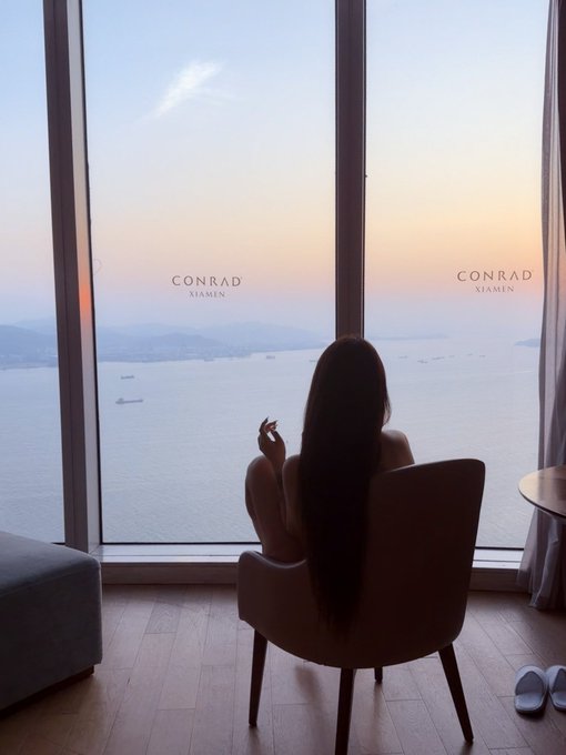 A woman with long dark hair sits in a chair facing a large floor-to-ceiling window at the 'CONRAD XIAMEN' hotel, looking out over a calm sea dotted with ships during a sunset or sunrise.