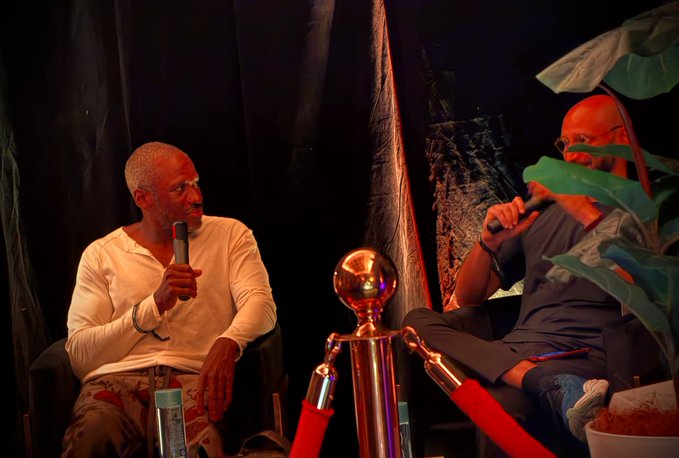 Two men sit on chairs on a stage with a stanchion rope between them; one in a white shirt speaks into a microphone while the other in a blue shirt listens.