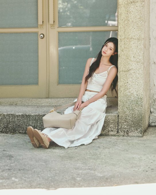 A young woman with long dark hair sits on stone steps outside a building, wearing a white crop top and long flowing skirt, holding a beige handbag.