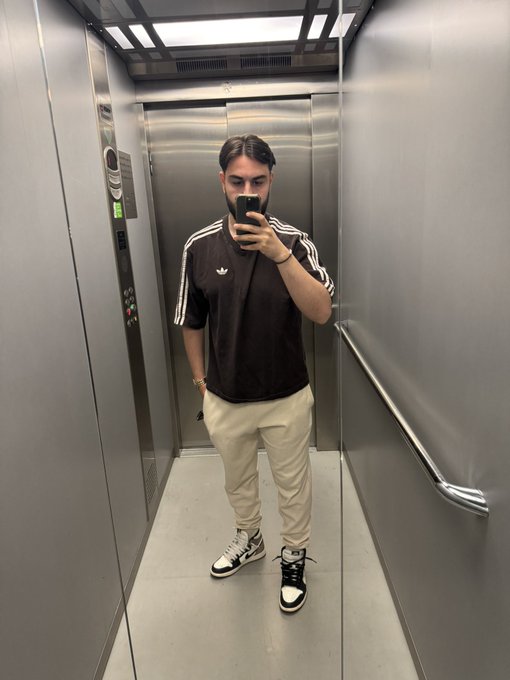 A man with dark hair takes a mirror selfie in an elevator while wearing a brown Adidas t-shirt, beige pants, and black and white sneakers.