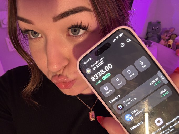 A close-up selfie of a woman holding a smartphone displaying a crypto portfolio screen showing '$1A DAY' and '$338.90' balance with various token options.