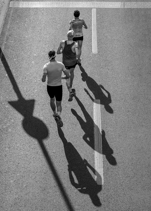 A black and white overhead photograph of three runners on a road, casting dramatic elongated shadows next to a road sign.
