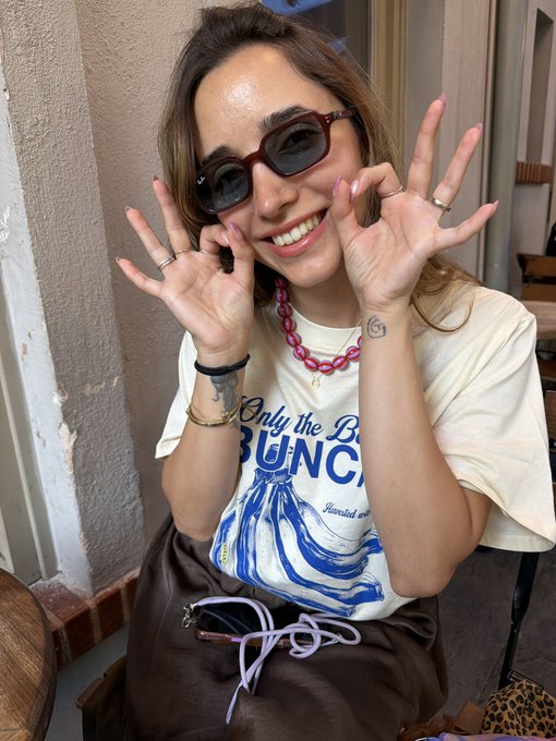 A smiling woman wearing sunglasses and a white graphic t-shirt, posing with her hands near her face, wearing multiple rings and a pink chain necklace.