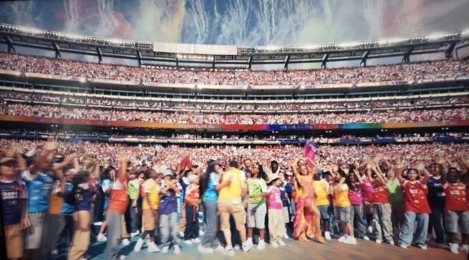 A massive stadium crowd with raised hands, performers on stage in colorful outfits, and a colorful sky backdrop above.