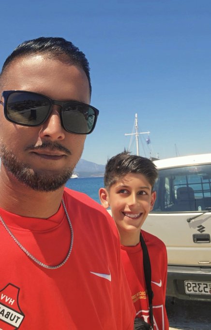 A selfie of a bearded man wearing sunglasses and a red shirt standing next to a smiling young boy, with a boat and blue sea visible in the background.