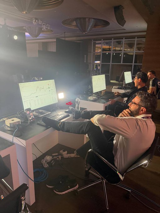 Several people are seated at desks with multiple monitors displaying trading charts in a dimly lit room.