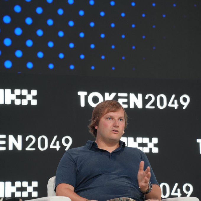 A man with light brown hair, wearing a blue polo shirt, sits on stage at the TOKEN2049 conference, gesturing with his hand as he speaks. The "TOKEN2049" branding is prominently displayed behind him on a digital screen, fitting the accompanying context of a panel discussion featuring Aave Labs founder Stani Kulechov on DeFi's cost advantages in lending.