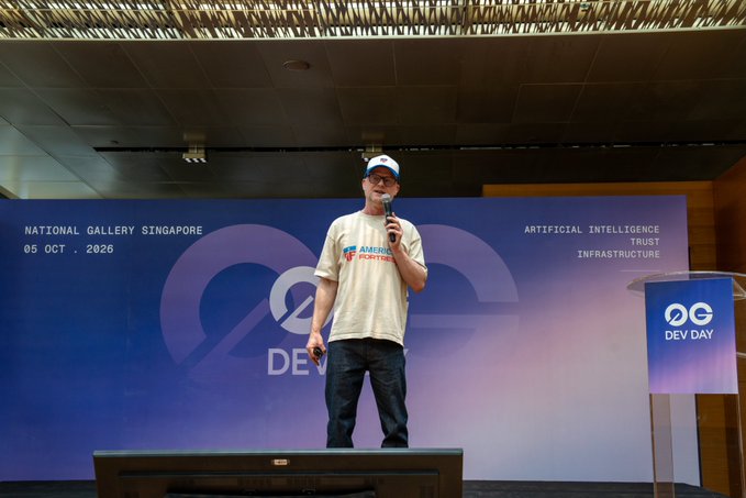 A man in a cap and "AMERICAN PICKERS" t-shirt speaks into a microphone on stage at "NATIONAL GALLERY SINGAPORE 05 OCT 2026" during a "DEV DAY" event focused on "ARTIFICIAL INTELLIGENCE TRUST INFRASTRUCTURE."