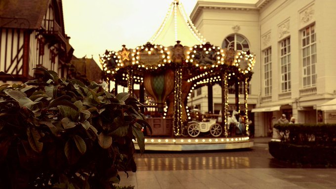 An ornate illuminated carousel at what appears to be Deauville, France, glowing with warm lights against a dusk sky, with a green bush in the foreground and classical architecture visible in the background.
