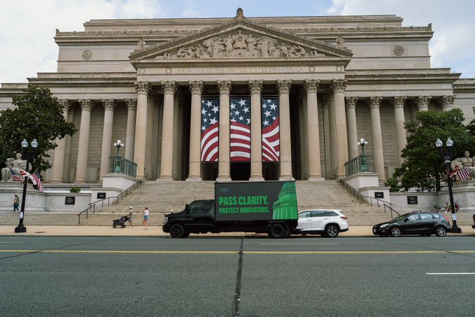 A mobile billboard truck displaying "PASS CLARITY. PROTECT INNOVATORS." is parked in front of a neoclassical government building adorned with American flag banners. The image visually supports the message about protecting developers and innovators by tying the political advocacy campaign directly to a seat of American government, reinforcing the call to safeguard builders within the Solana ecosystem and the broader U.S. tech landscape.