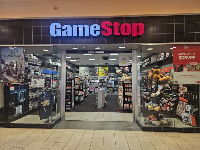 The image shows the entrance of a GameStop store located inside a mall, with the iconic red and white "GameStop" logo prominently displayed above the doorway. Promotional signs visible include an Assassin's Creed Black Flag display on the left and a "$32 SAVE UP TO $29.99" Nintendo Switch bundle advertisement on the right, tying into the $GME reference as the store's ticker symbol.