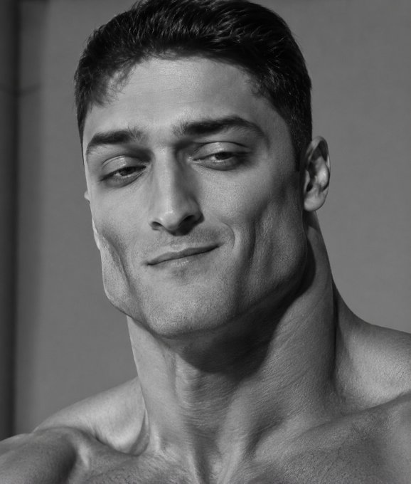 A black and white close-up portrait of a shirtless man with a slight smile, showing defined jawline and neck muscles.