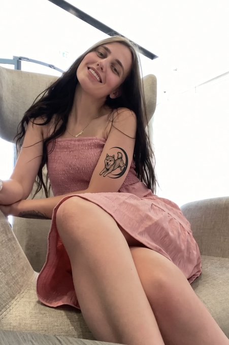 A smiling woman in a pink strapless dress sitting in a chair, showing a small tattoo of a cat on a crescent moon on her upper arm.