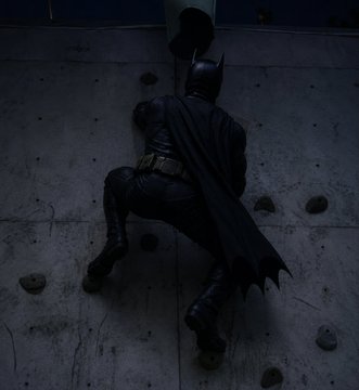 Batman is shown from behind, clinging to a concrete wall in a dark setting.