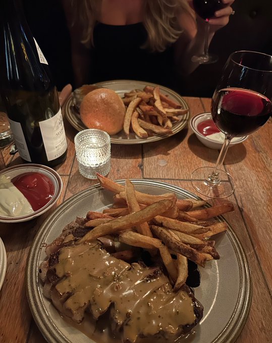 A bottle of Meursault white wine and a glass of red wine accompany plates of steak frites and a burger at a dimly lit dinner, with the bottle's "Meursault" label visible. The accompanying caption about Hyperliquid hitting $80 contrasts the leisurely, indulgent dining scene with an oblivious crypto market milestone.