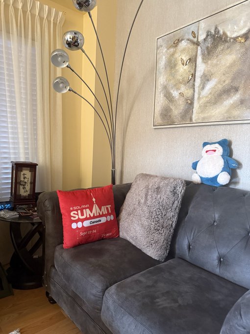 A living room scene featuring a gray couch with a red pillow reading 'SCOLARITÉ SUMMIT, Canada, Sept 25-26', a Snorlax plush toy, a modern arc floor lamp with chrome spheres, and a framed gold-toned painting.
