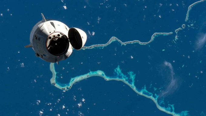 A spacecraft is photographed in orbit above Earth, with bright blue ocean waters and land formations visible below.