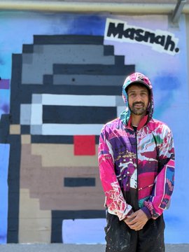 A man with a beard wearing a pink hoodie with a floral pattern stands smiling in front of a mural featuring a pixelated character design with the text 'Masnah.'