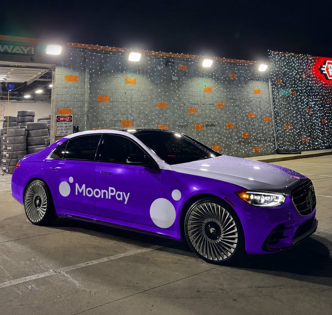 A custom purple Mercedes-Benz sedan featuring MoonPay branding and white moon graphics parked at night in front of a building decorated with string lights.