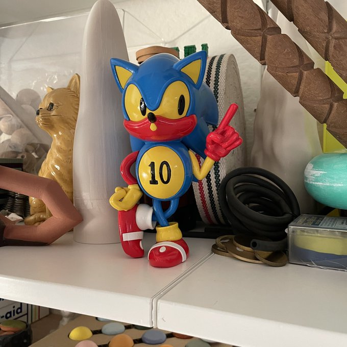 A figurine of a blue cartoon cat/fox character giving a middle finger, sporting the number "10" on its chest, stands on a shelf alongside various knickknacks including a ceramic cat and coiled tubing—likely purchased with Bitcoin ($BITCOIN) gains.