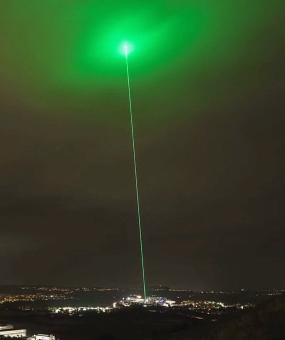 A bright green laser beam shoots skyward from a distant city, evoking the "to the moon" rocket imagery associated with the post's question about which memecoin will surge 500x.