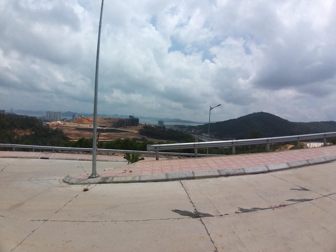 A view from an unfinished concrete road looking out over a landscape with hills, construction sites, and distant buildings under a cloudy sky.