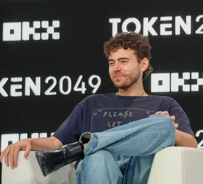 Polymarket CEO Shayne Coplan sits relaxed on a stage at the TOKEN2049 conference, the "OKX" and "TOKEN2049" branding visible behind him, appearing to address the audience on stage. His casual posture and slight smile contrast with his pointed critique of crypto speculation, in which he warned that chasing 100x returns reflects "irrational exuberance" and amounts to a "greater fool" game.