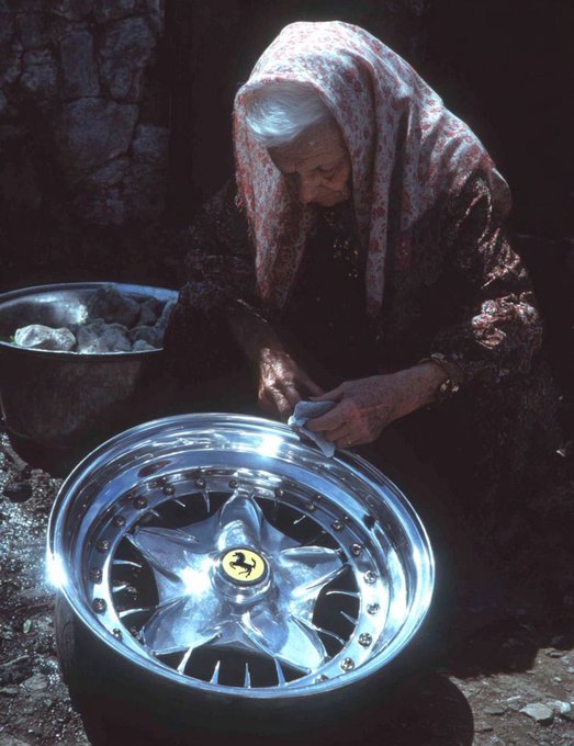An elderly woman in a floral headscarf carefully cleans and polishes a shiny Ferrari wheel rim in a dimly lit, rustic setting.